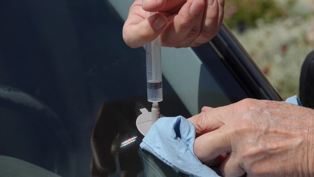 Close up of a man's hands repairing small impact hole in car windshield with DIY kit